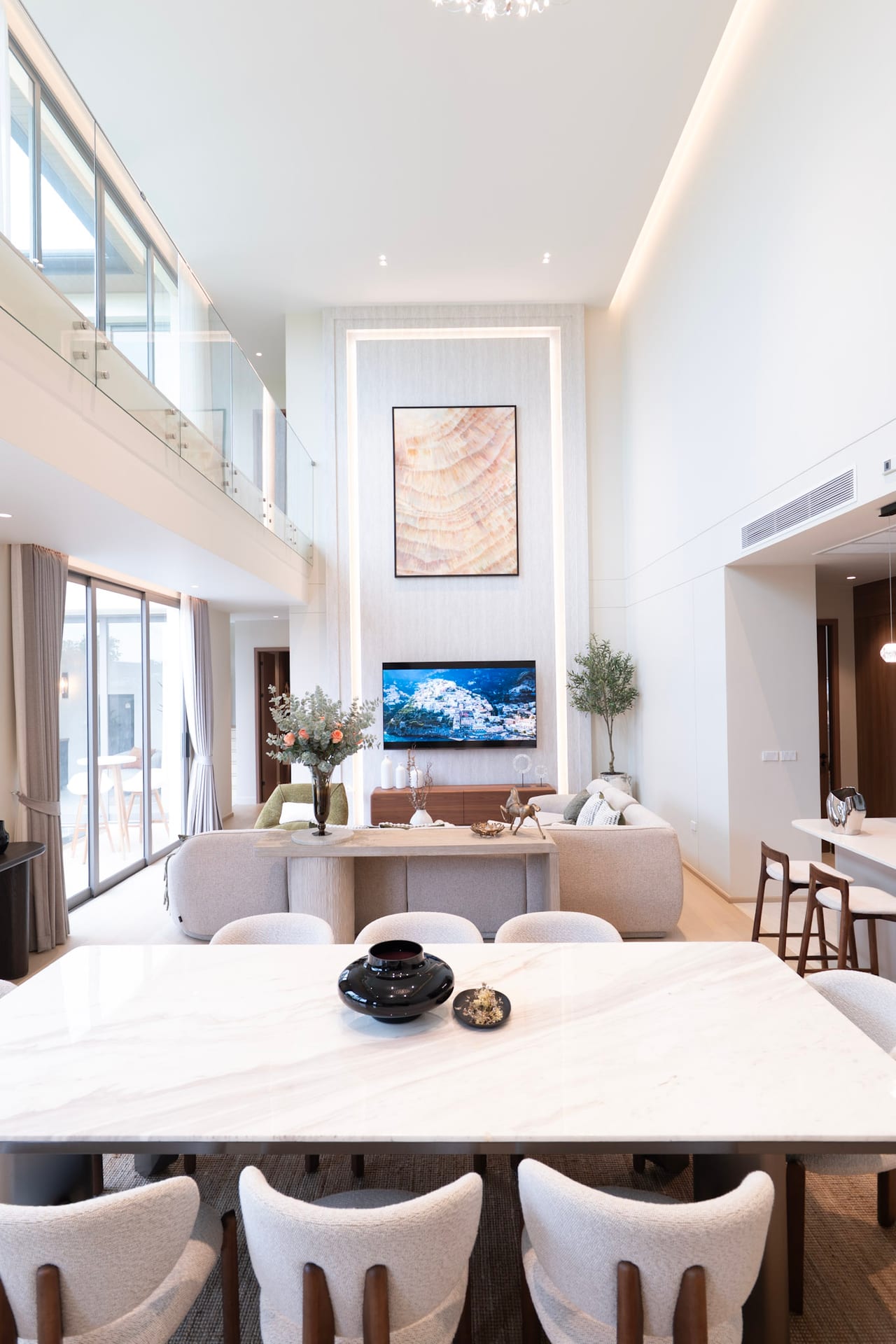 Bright open living room with a marble dining table in the foreground and beige chairs; wall-mounted TV and framed abstract artwork on a tall recessed panel in the center.