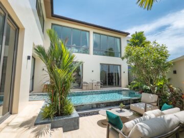 Modern two-story home courtyard with large glass doors, pool, and tropical plants under a blue sky