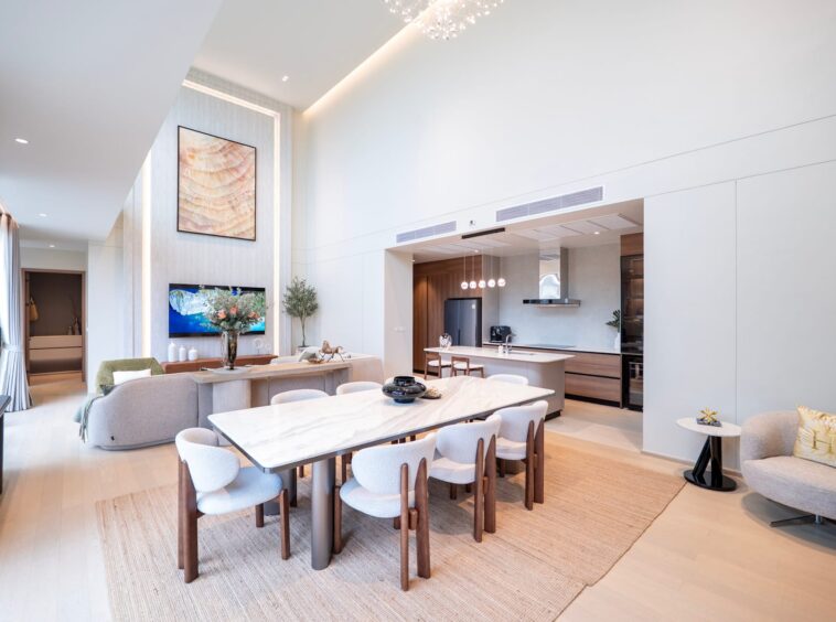 Bright open-plan living, dining, and kitchen area with marble dining tables, light wood floors, and soft beige seating.