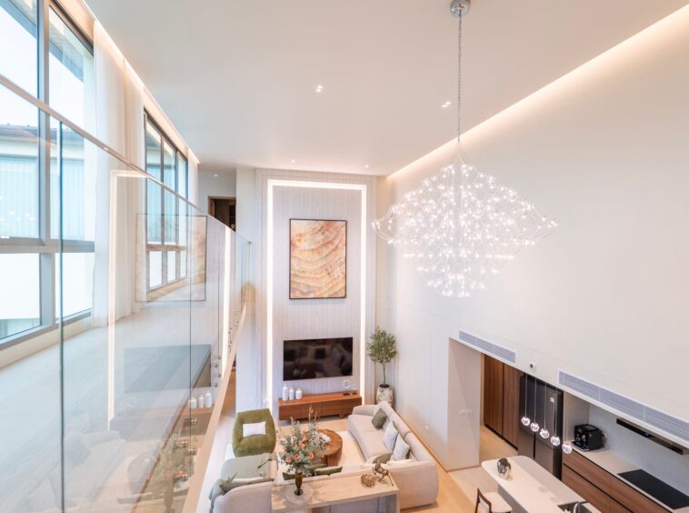 Bright, modern living room with a large crystal chandelier, glass railing, and a wall-mounted TV centerpiece.