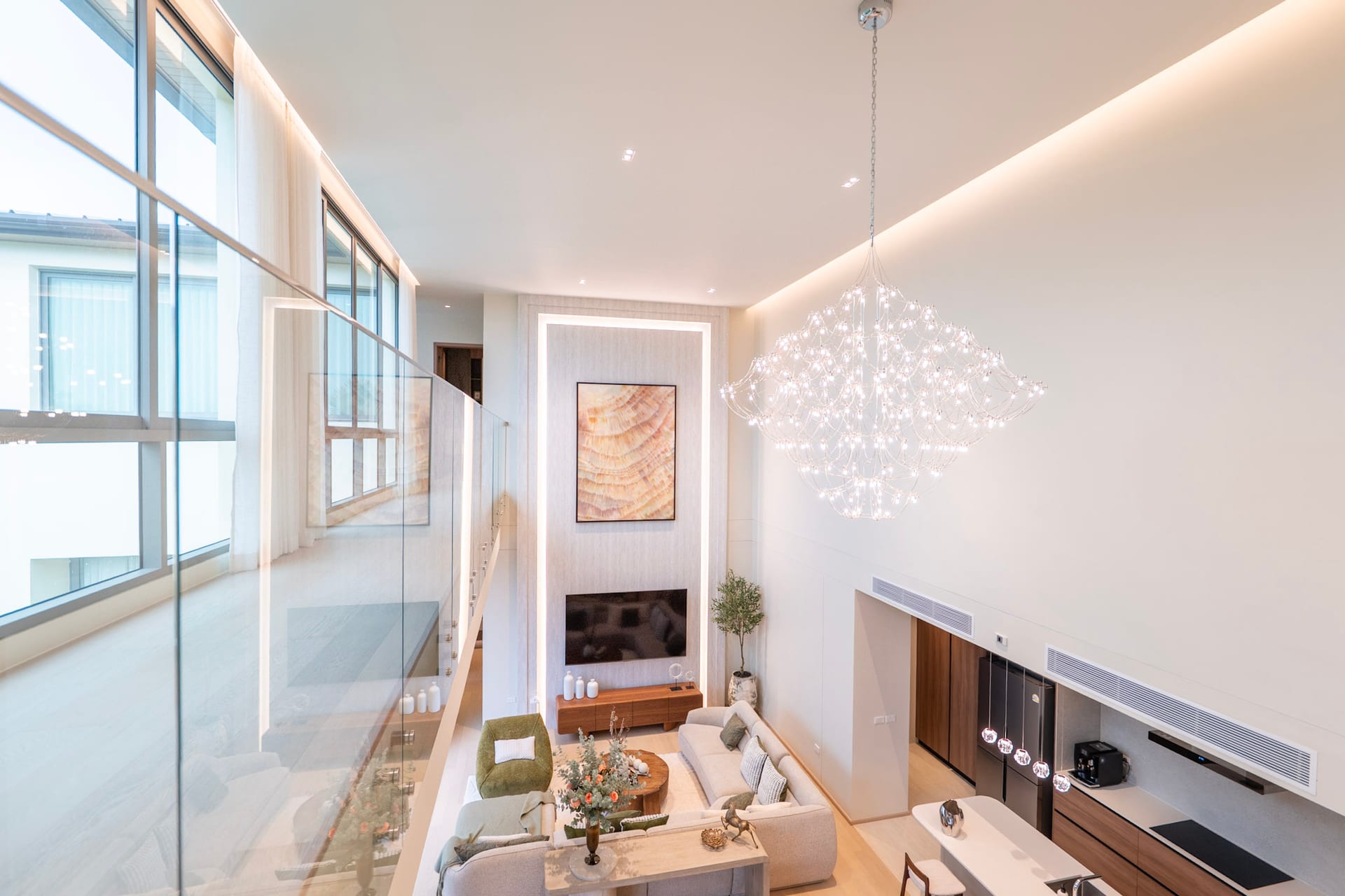 Bright, modern living room with a large crystal chandelier, glass railing, and a wall-mounted TV centerpiece.