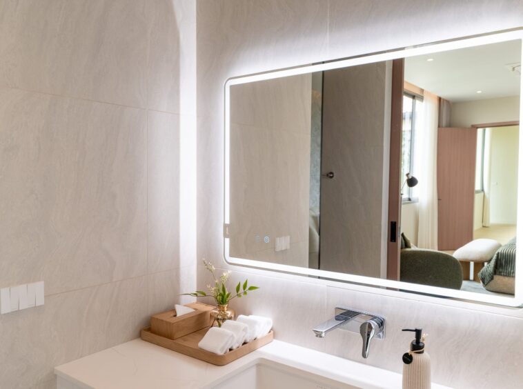 Illuminated rectangular mirror above a white bathroom sink, beige marble-like tiles, with a wooden tray of towels and a small plant on the counter; reflection shows a living area with sofa and window.