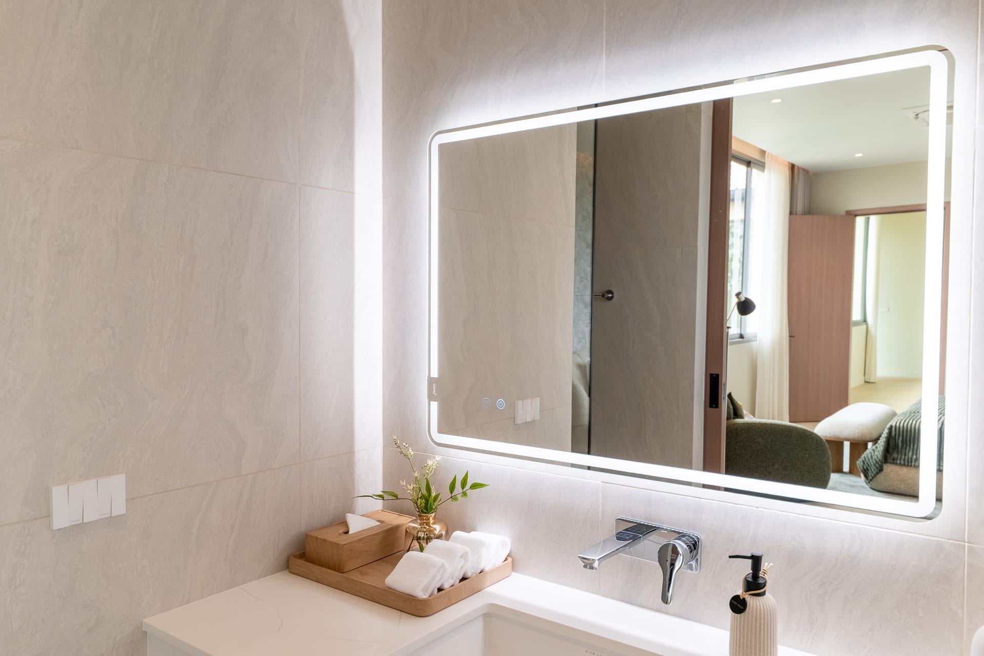 Illuminated rectangular mirror above a white bathroom sink, beige marble-like tiles, with a wooden tray of towels and a small plant on the counter; reflection shows a living area with sofa and window.