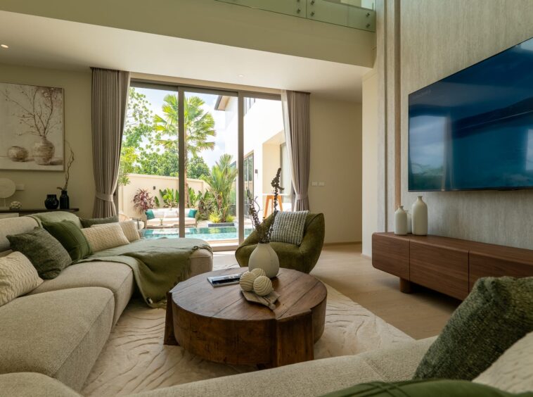 Bright modern living room with a beige sectional sofa, round wooden coffee table, and large sliding glass doors opening to a tropical outdoor patio and pool area