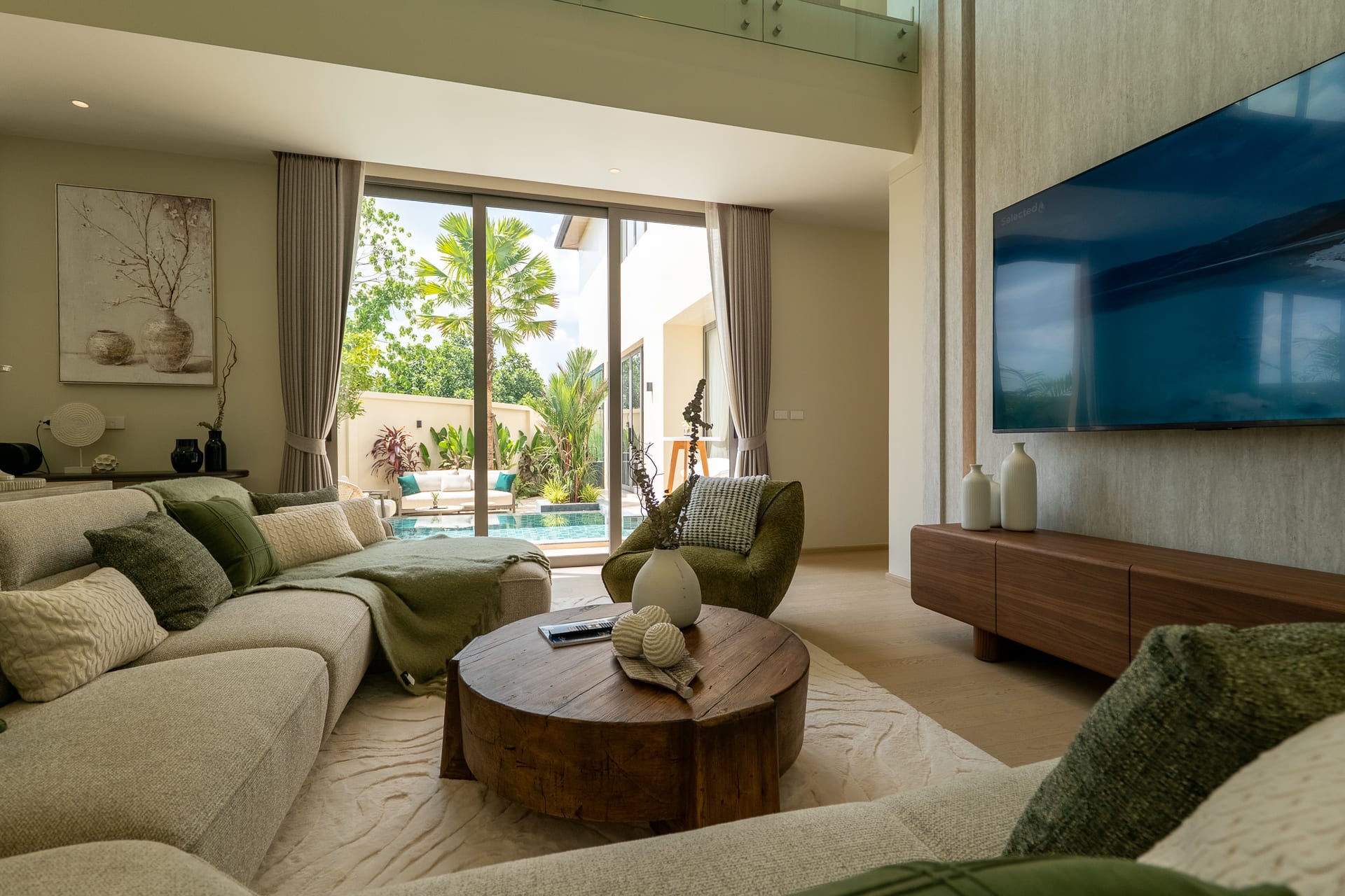 Bright modern living room with a beige sectional sofa, round wooden coffee table, and large sliding glass doors opening to a tropical outdoor patio and pool area