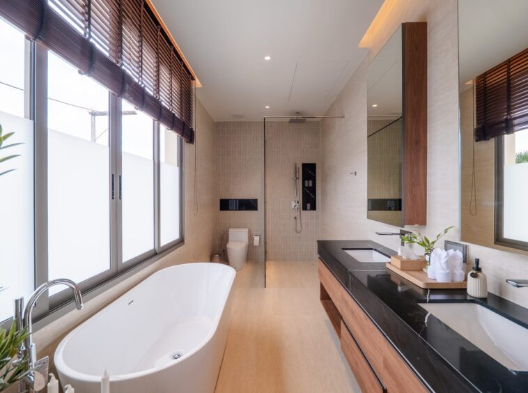 Bright contemporary bathroom with a freestanding white tub by large frosted windows, a glass-enclosed shower, and a long black marble vanity top with wood drawers.