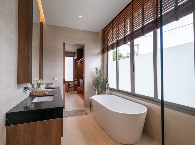 Modern bathroom with a white freestanding tub by large windows with wooden blinds on a bright, beige-tiled wall.