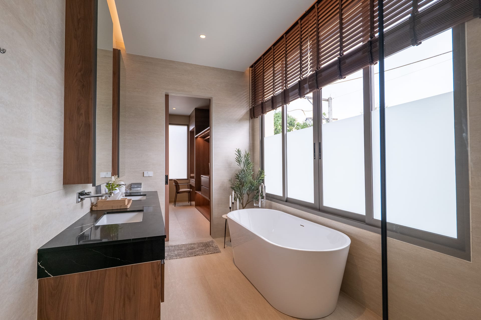 Modern bathroom with a white freestanding tub by large windows with wooden blinds on a bright, beige-tiled wall.