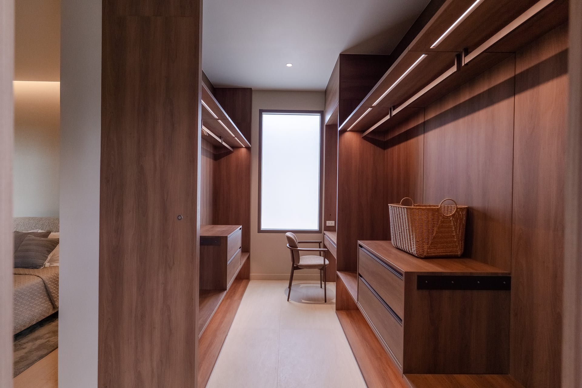 Wood-paneled walk-in closet with built-in storage on both sides, a central window, and a wicker basket on a dresser/console.