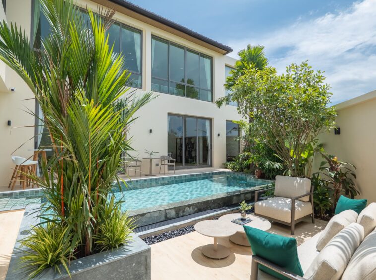 Modern two-story home with large glass windows, beige walls, and a rectangular tiled pool in a sunny courtyard. lush tropical plants frame the scene.