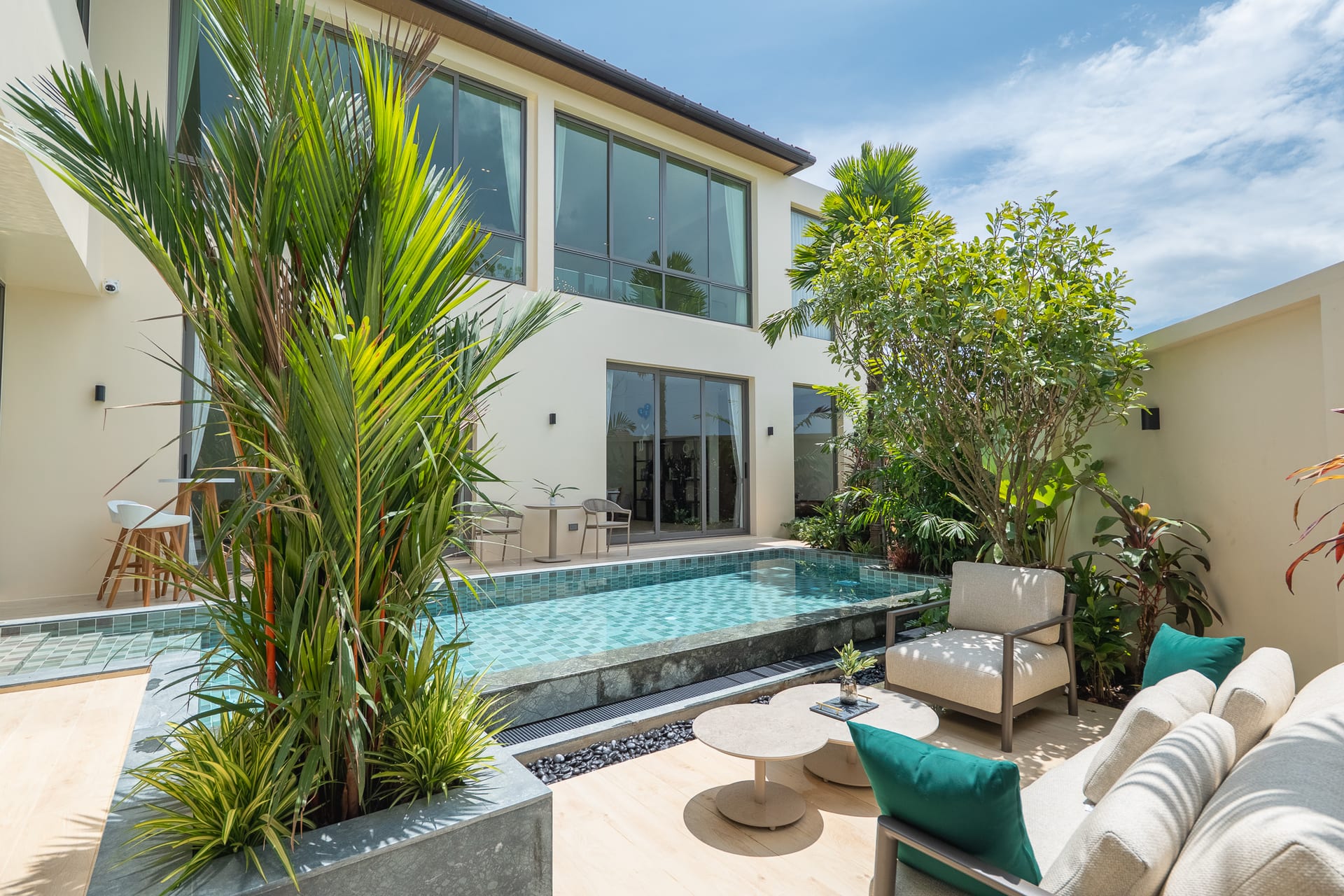 Modern two-story home with large glass windows, beige walls, and a rectangular tiled pool in a sunny courtyard. lush tropical plants frame the scene.