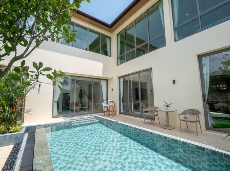 Modern house courtyard with a turquoise-tiled pool, glass sliding doors, and beige exterior walls surrounded by greenery.