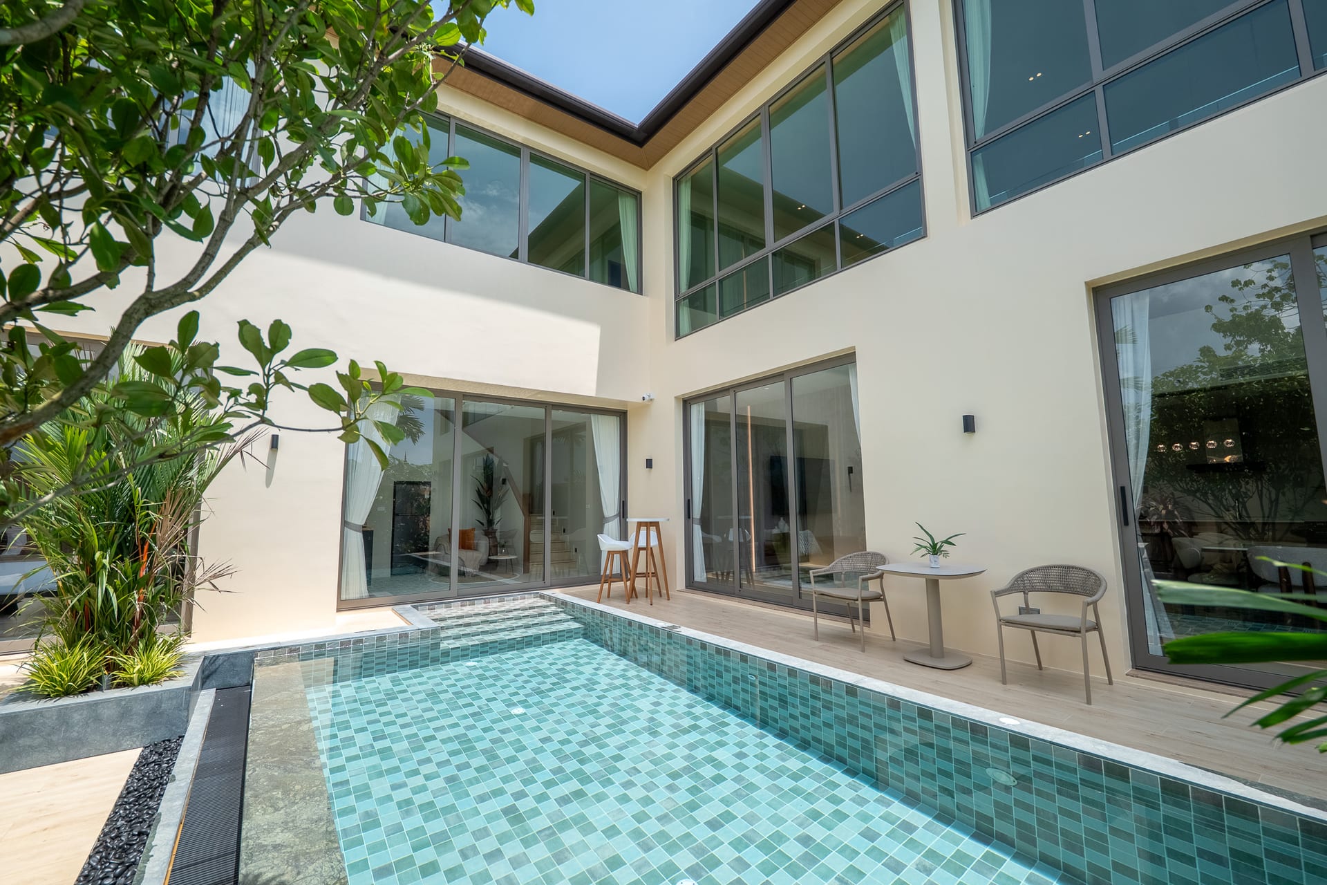 Modern house courtyard with a turquoise-tiled pool, glass sliding doors, and beige exterior walls surrounded by greenery.