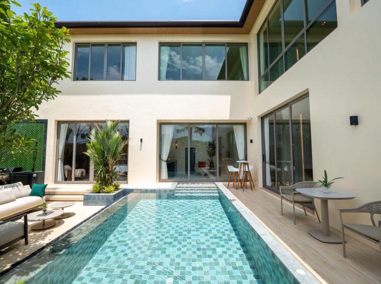 Contemporary two-story house with large glass doors, a turquoise tiled pool, and outdoor seating on a wooden deck.