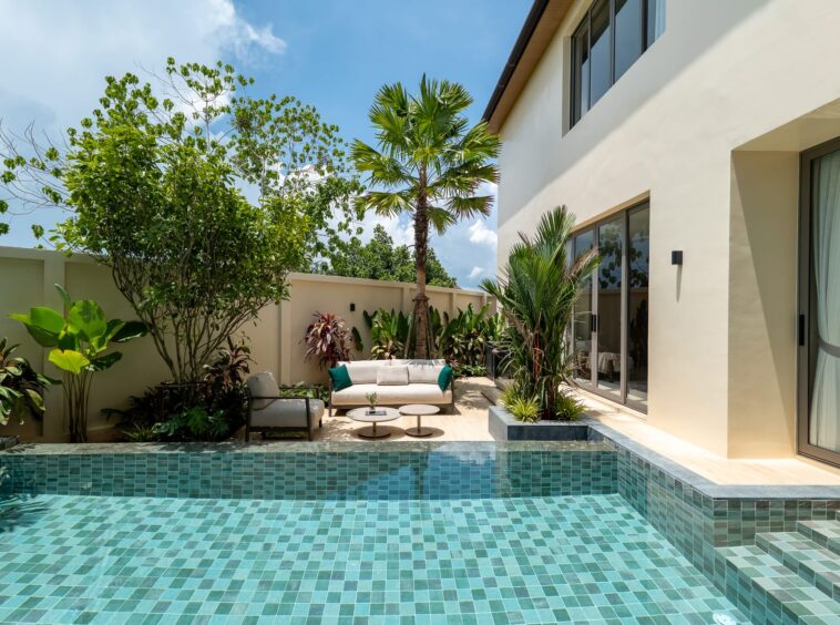 Pool area with turquoise tiled water and stepped edge beside a modern beige house and glass doors, surrounded by tropical plants and seating.