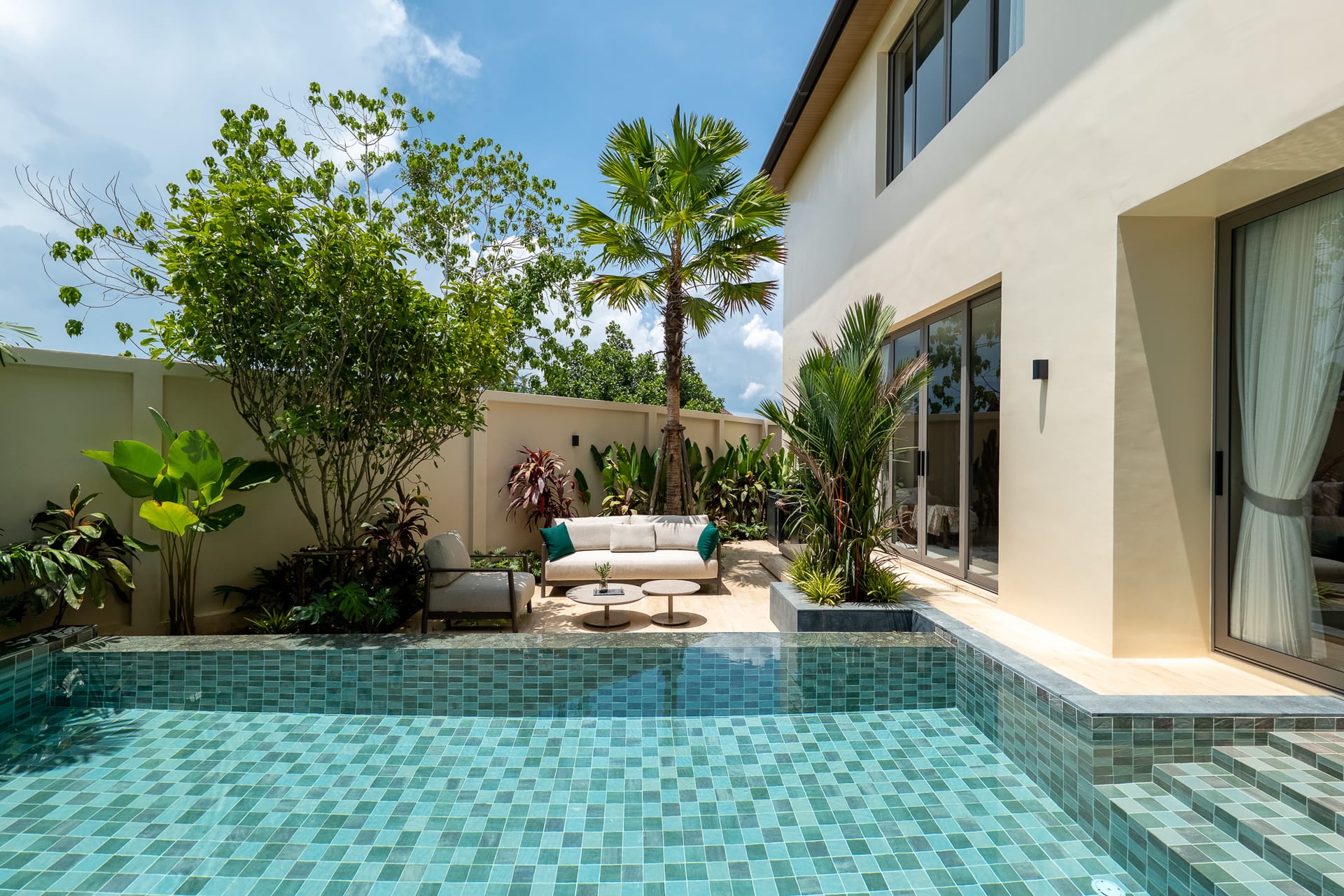 Pool area with turquoise tiled water and stepped edge beside a modern beige house and glass doors, surrounded by tropical plants and seating.