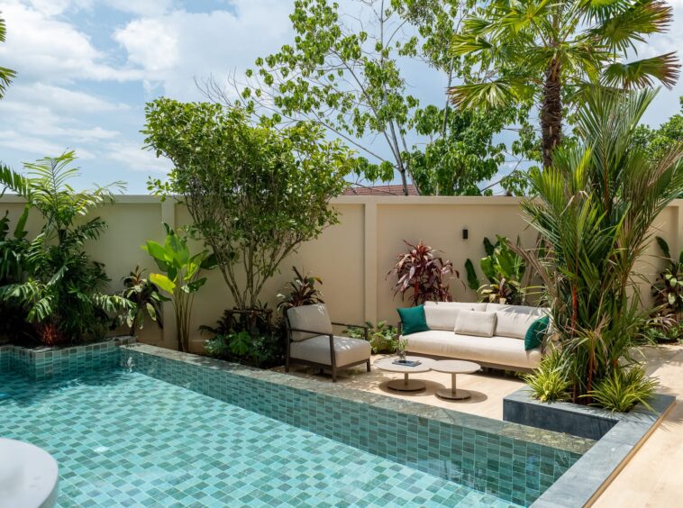 Backyard pool with turquoise-tiled water, beige seating area, and lush tropical plants against a beige wall.
