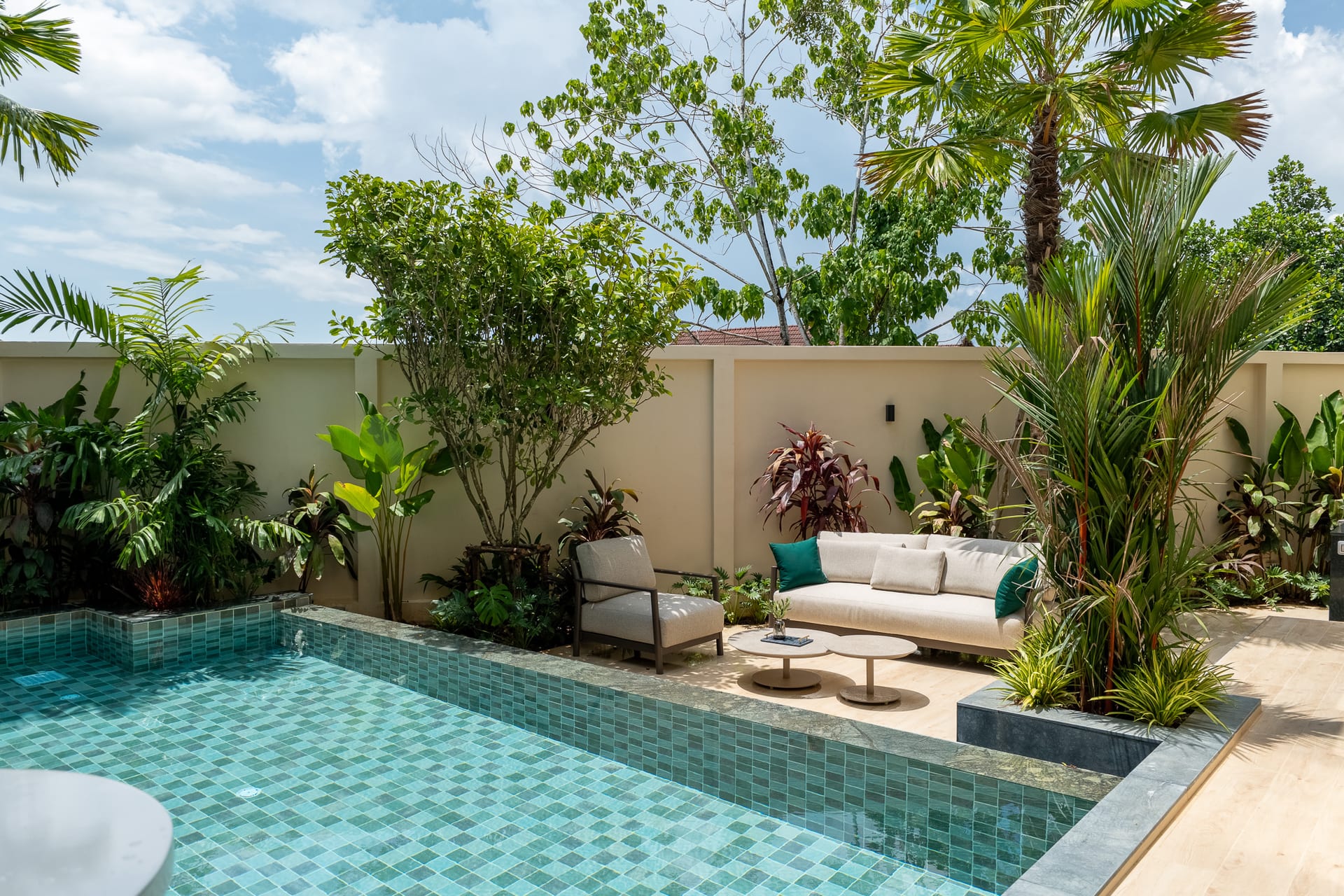 Backyard pool with turquoise-tiled water, beige seating area, and lush tropical plants against a beige wall.