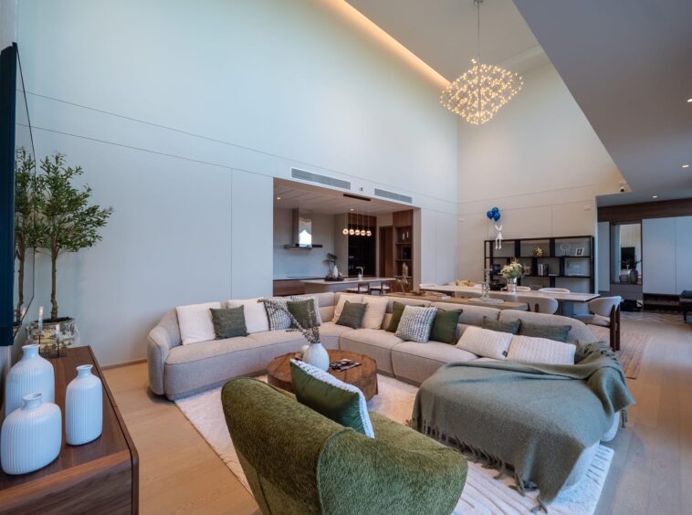 Open-concept living room with a large L-shaped beige sofa, green cushions, round wooden coffee table, tall plants, and a sculptural chandelier overhead.