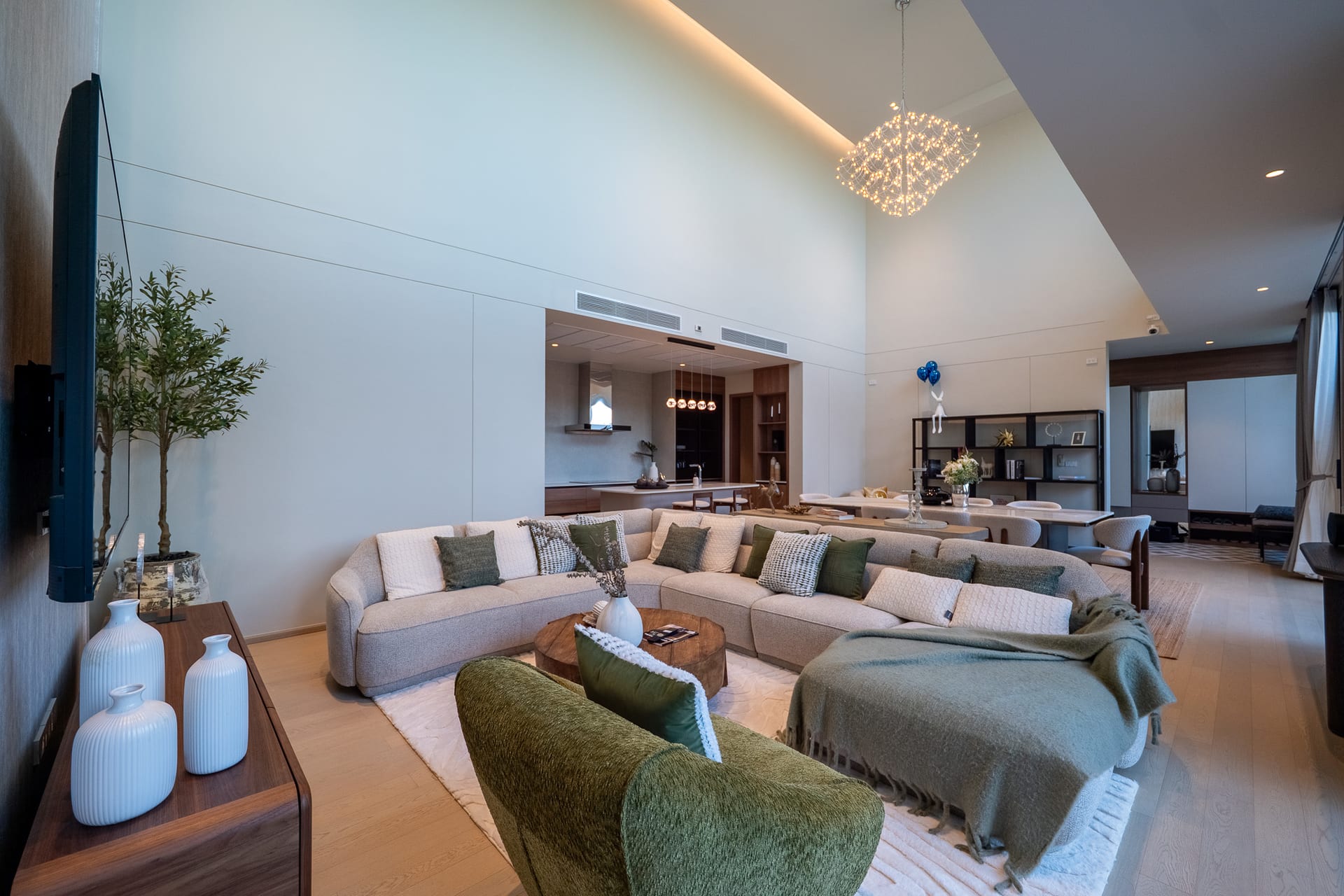 Open-concept living room with a large L-shaped beige sofa, green cushions, round wooden coffee table, tall plants, and a sculptural chandelier overhead.