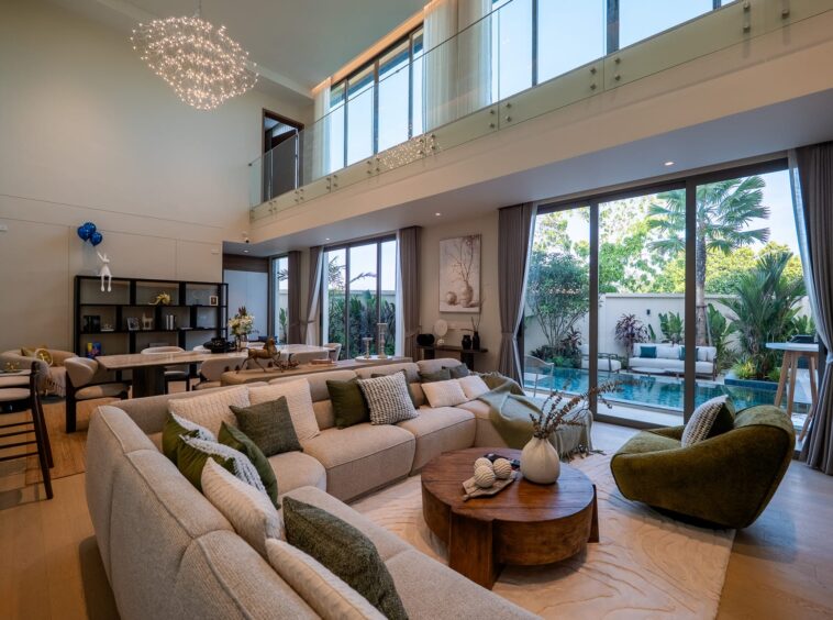 Spacious two-story living room with beige sofa seating, green cushions, and two round wooden coffee tables, overlooking a glass railing and large glass doors to the outdoor pool area.