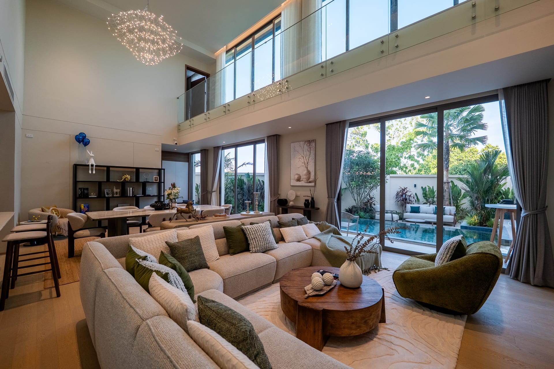 Spacious two-story living room with beige sofa seating, green cushions, and two round wooden coffee tables, overlooking a glass railing and large glass doors to the outdoor pool area.