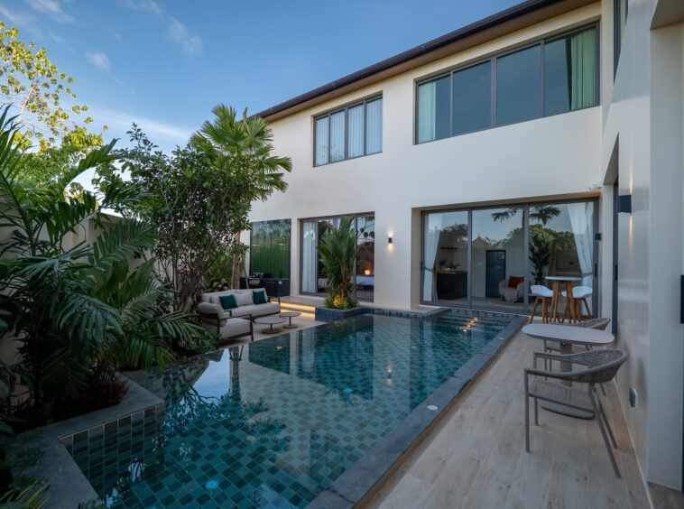 Modern two-story home with a glass-walled back, poolside seating, and lush tropical plants.