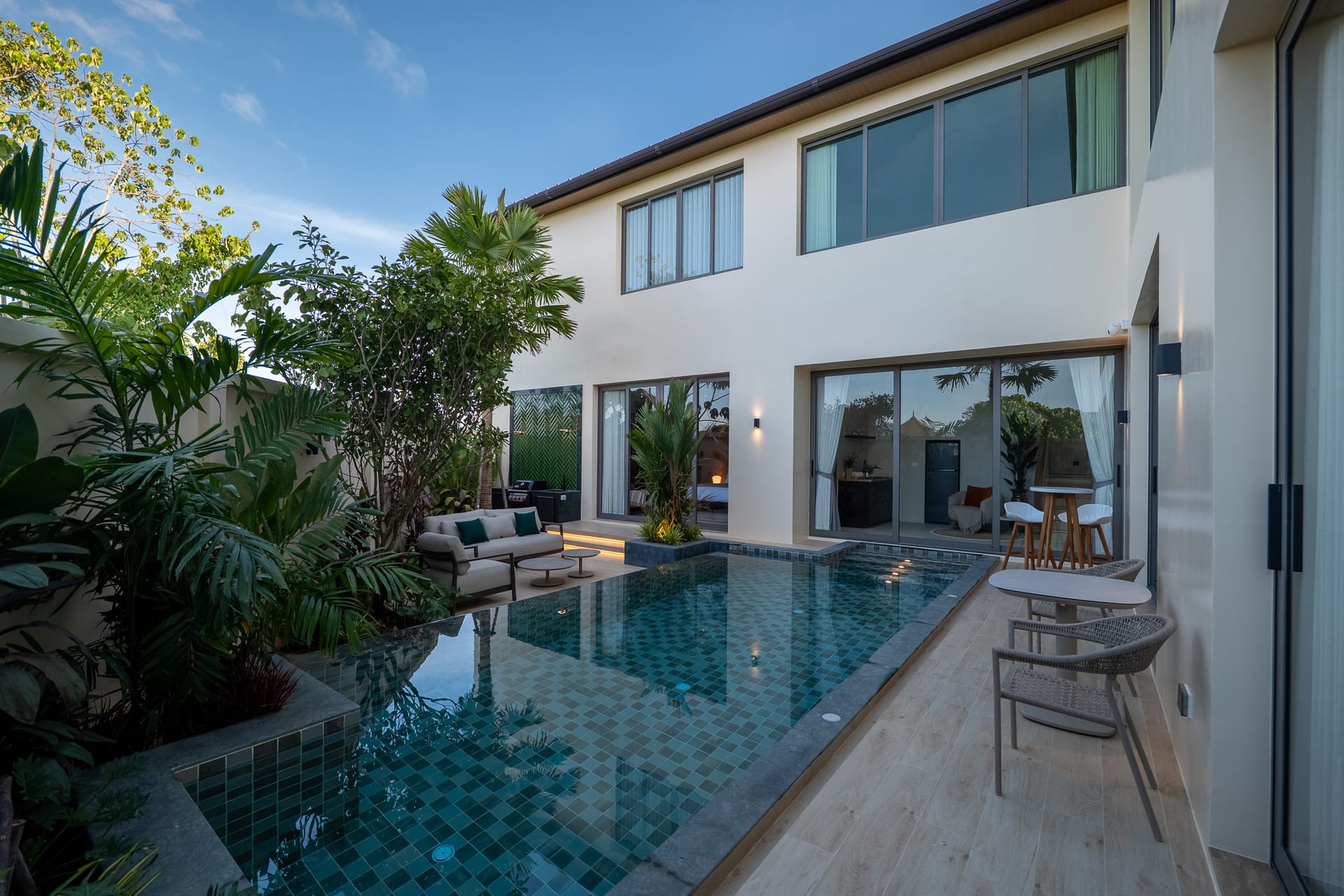 Modern two-story home with a glass-walled back, poolside seating, and lush tropical plants.