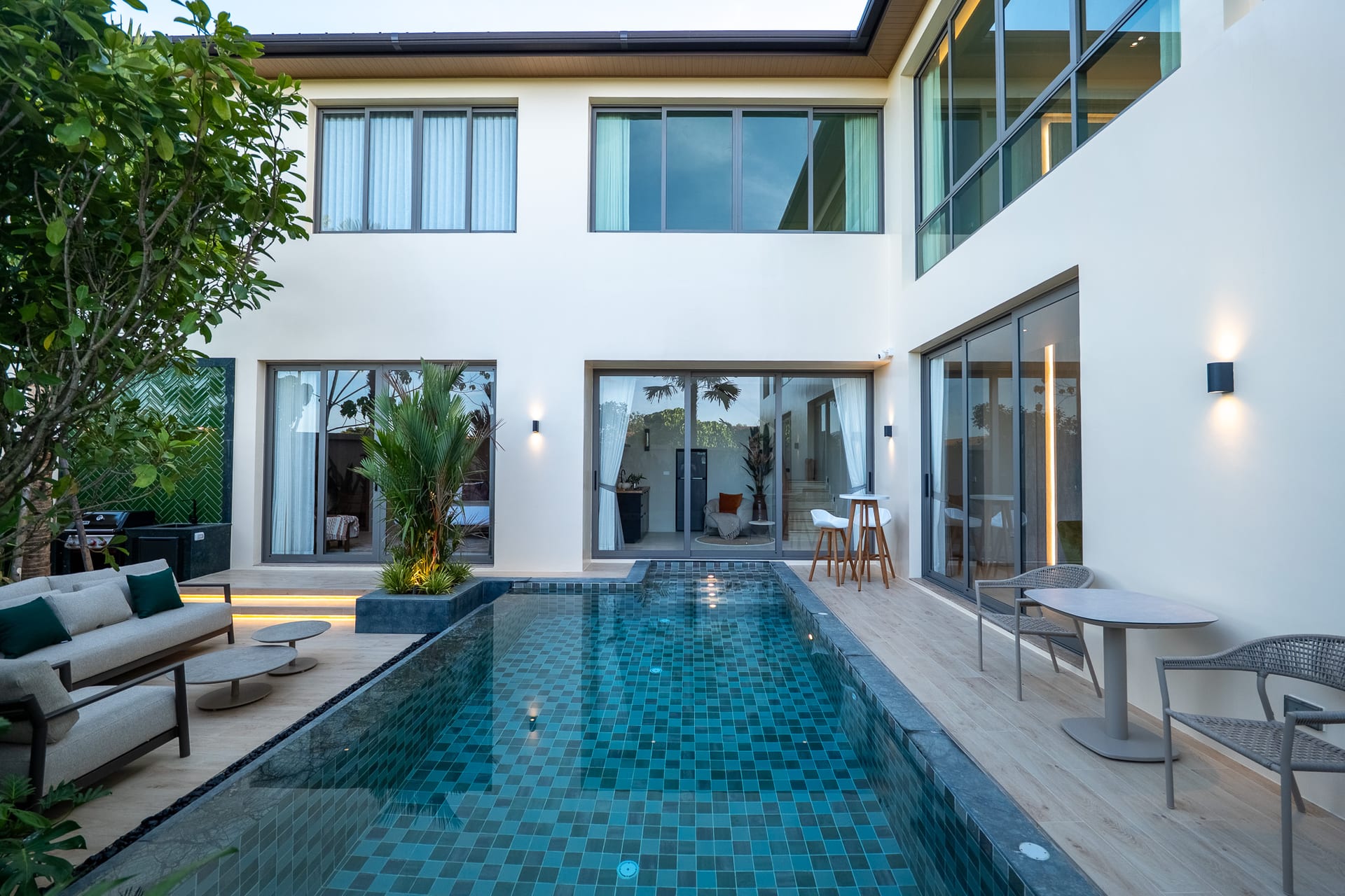 Modern courtyard pool with blue mosaic tiles, lounge seating, and glass doors opening to indoors
