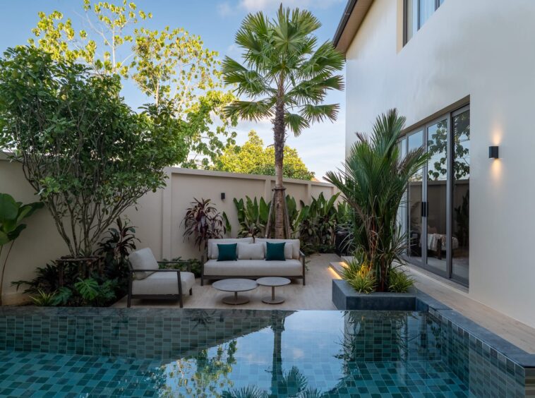 Modern backyard courtyard with a rectangular turquoise-tiled pool, beige seating and tropical plants along a cream wall.