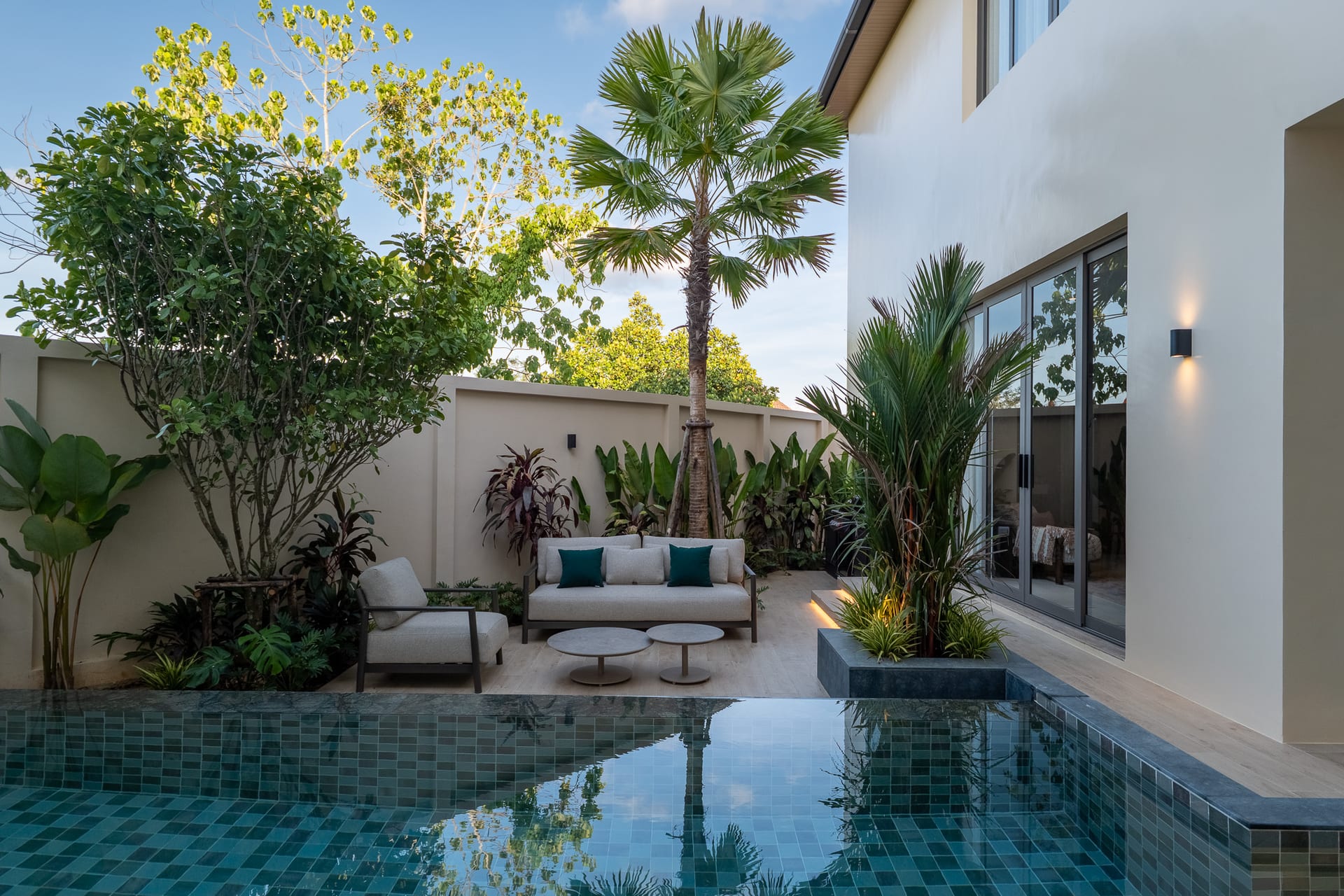 Modern backyard courtyard with a rectangular turquoise-tiled pool, beige seating and tropical plants along a cream wall.