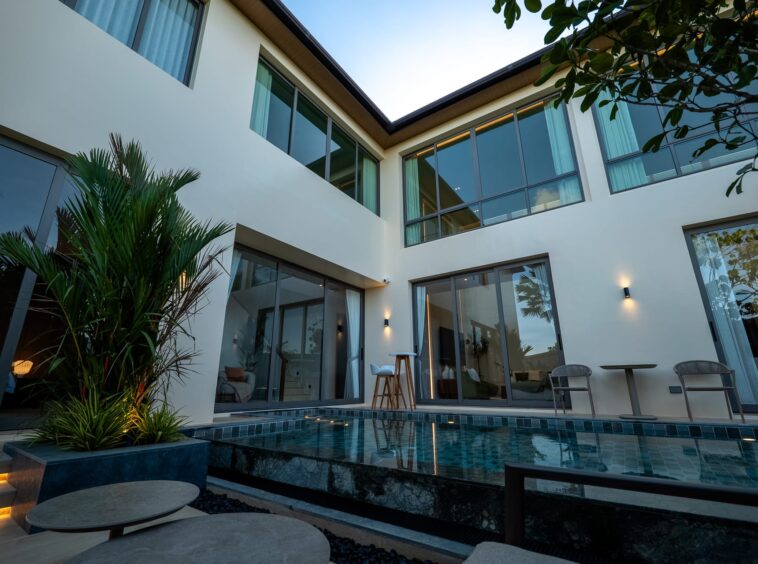 Modern glass-walled courtyard of a white two-story home with a rectangular pool and outdoor seating, sunset lighting, and lush plants
