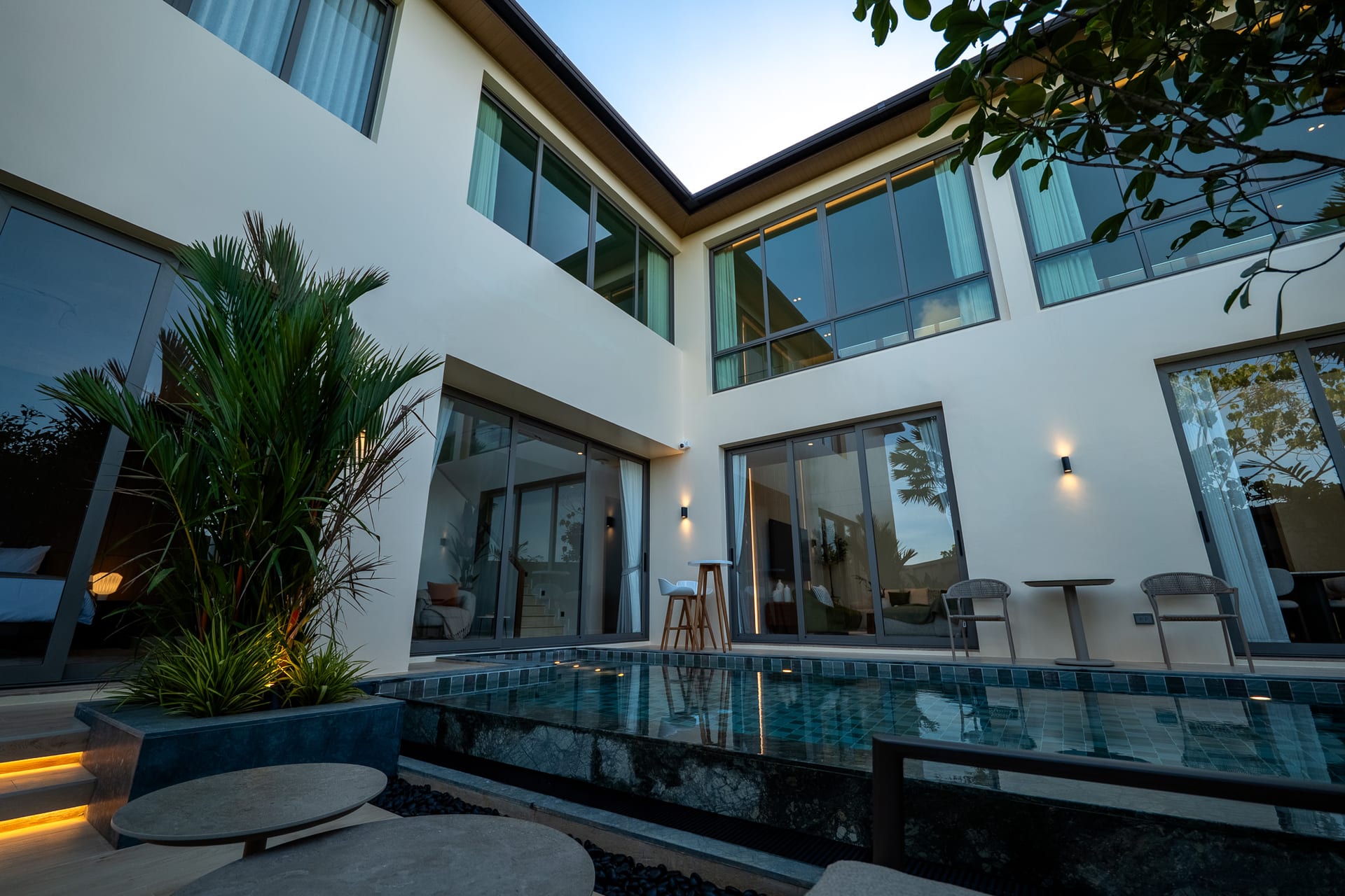 Modern glass-walled courtyard of a white two-story home with a rectangular pool and outdoor seating, sunset lighting, and lush plants