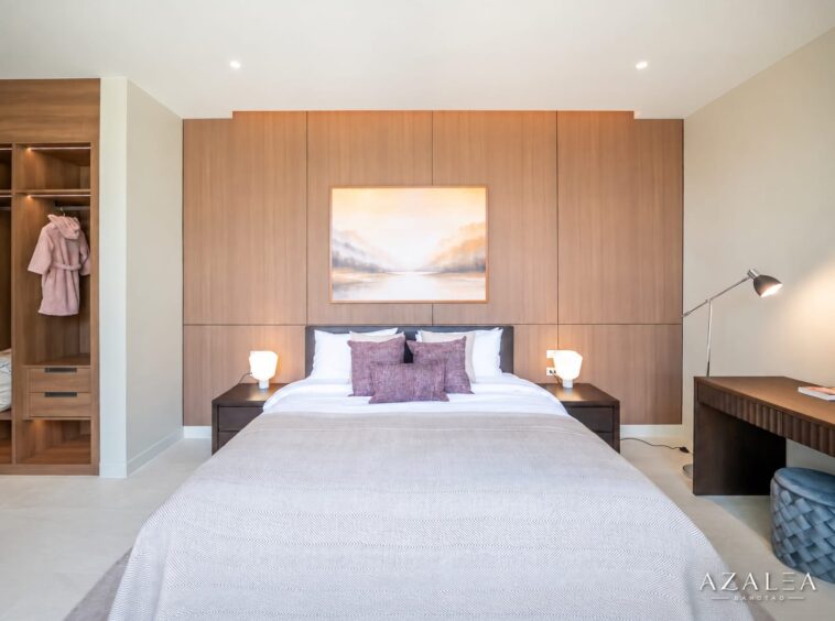 Modern bedroom with a large wood-paneled wall behind a king bed, framed landscape painting, and two lit bedside lamps on dark wood nightstands on either side.