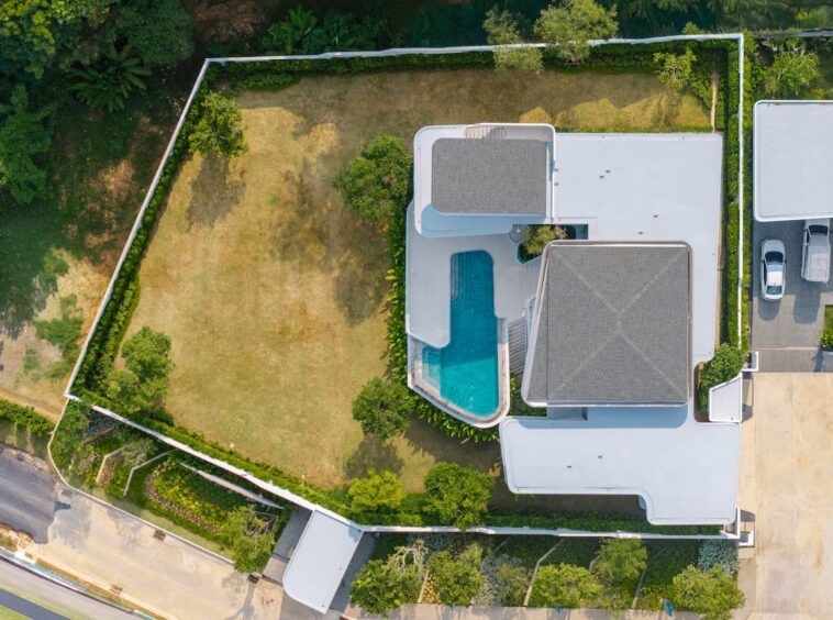 Aerial view of a modern house with a curved-in pool and white rooftop structures surrounded by a fenced yard.