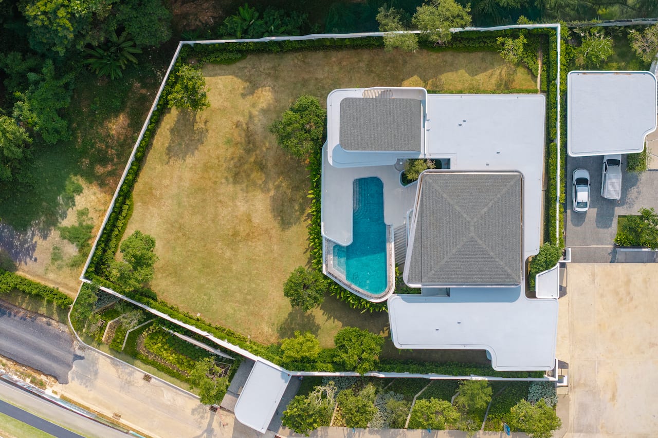 Aerial view of a modern house with a curved-in pool and white rooftop structures surrounded by a fenced yard.