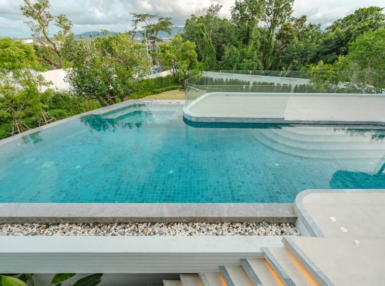 Modern outdoor infinity pool with turquoise water, glass railing, and lush trees in the background