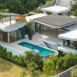 Aerial view of a modern white villa with glass balconies, a curved pool, and manicured greenery nearby.