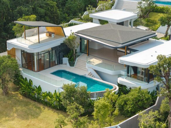 Aerial view of a modern white villa with glass balconies, a curved pool, and manicured greenery nearby.