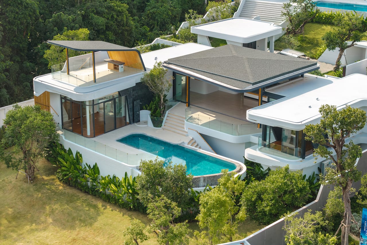 Aerial view of a modern white villa with glass balconies, a curved pool, and manicured greenery nearby.