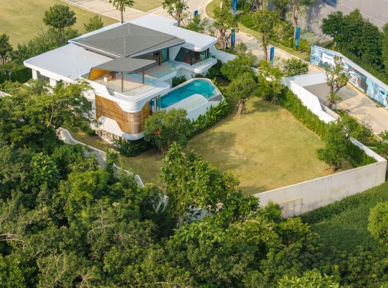 Aerial view of a modern white villa with multiple terraces, shaded areas, and a curved swimming pool surrounded by trees and greenery, from above.