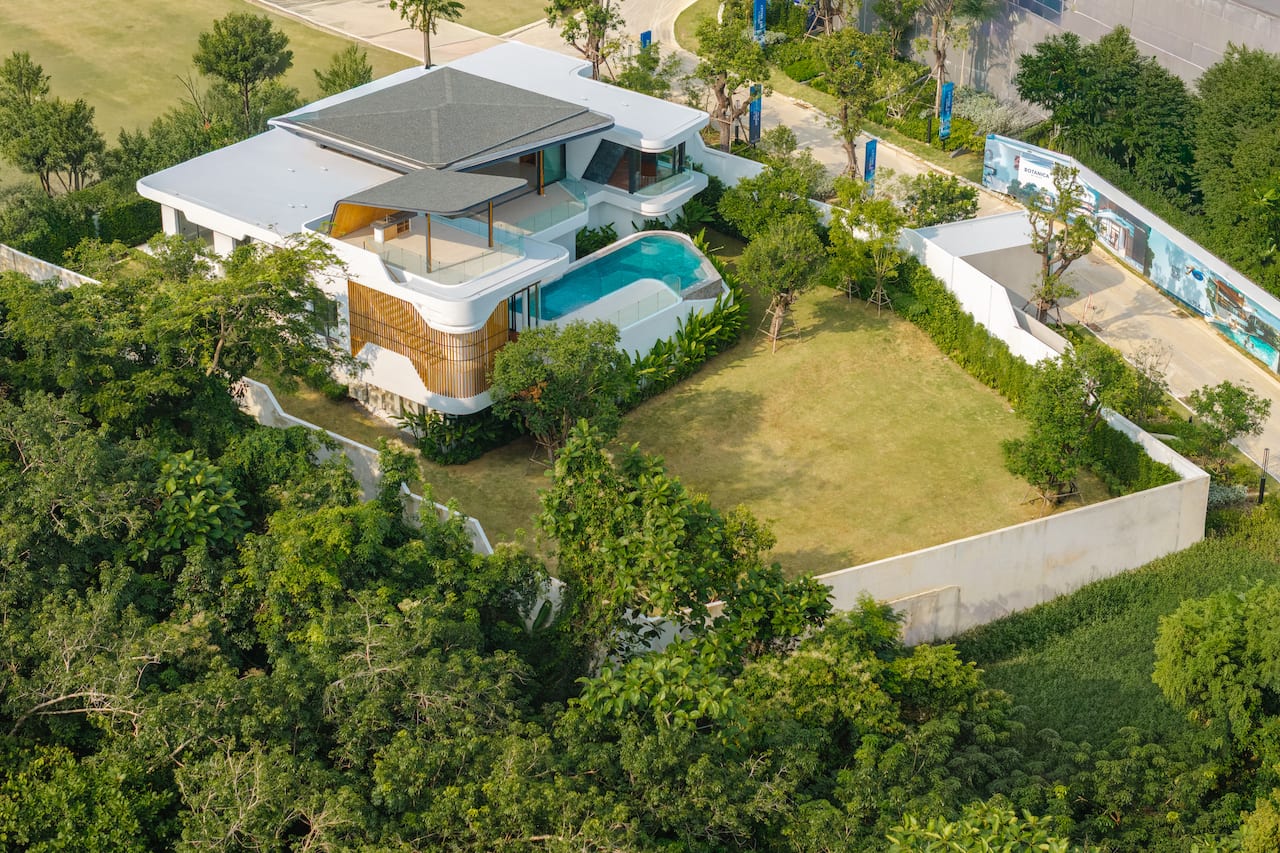 Aerial view of a modern white villa with multiple terraces, shaded areas, and a curved swimming pool surrounded by trees and greenery, from above.