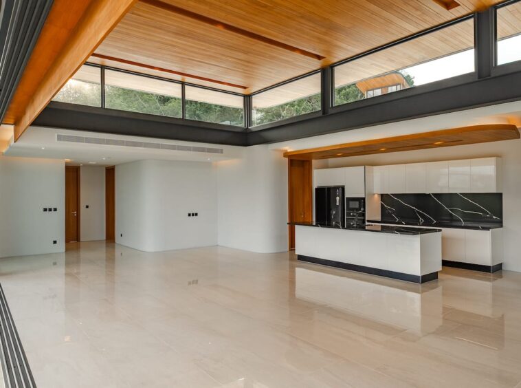Bright open-plan kitchen and living area with a white island, black marble backsplash, and glossy beige tile floors under a timber ceiling and large glass walls.