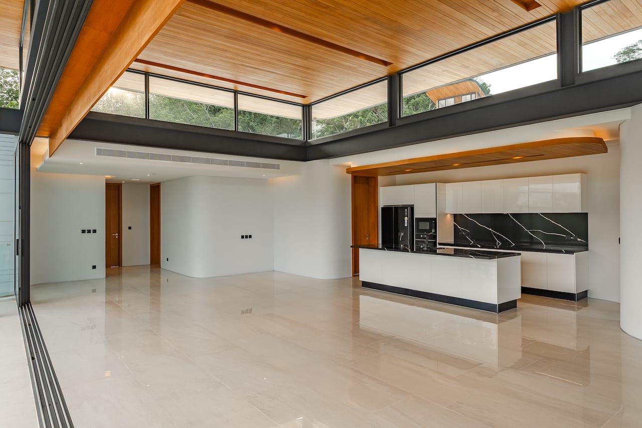 Bright open-plan kitchen and living area with a white island, black marble backsplash, and glossy beige tile floors under a timber ceiling and large glass walls.