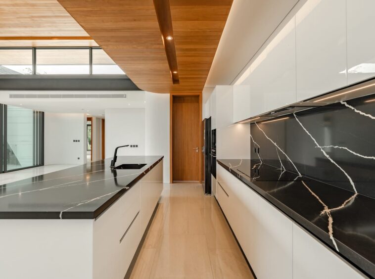 Modern white kitchen with black marble countertops, a long island sink, and wooden ceiling beams.