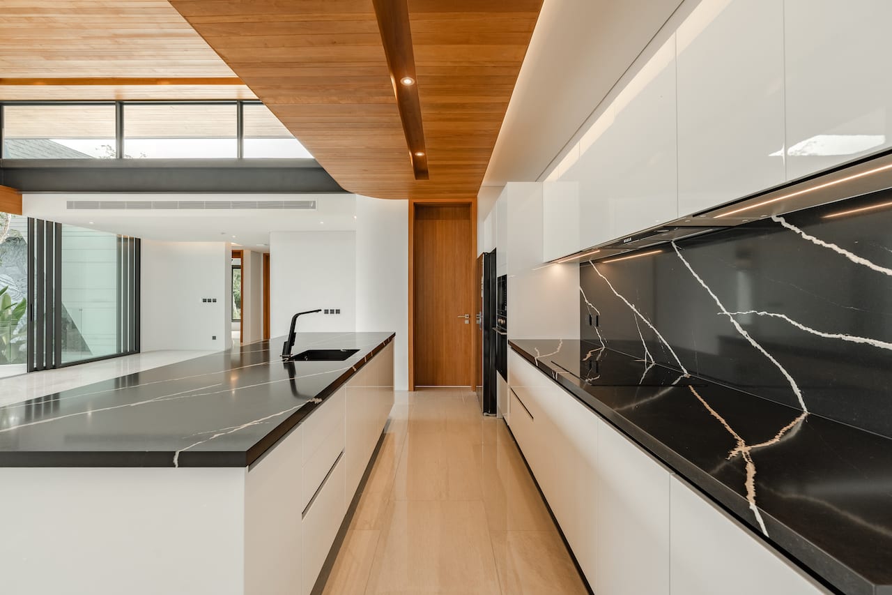 Modern white kitchen with black marble countertops, a long island sink, and wooden ceiling beams.