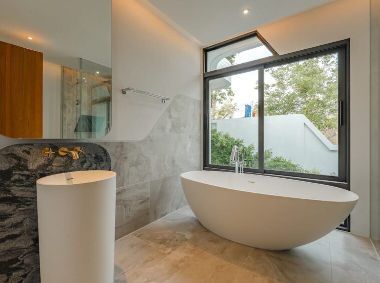 Modern bathroom with a freestanding oval tub by a large window and marble-tiled walls and floor