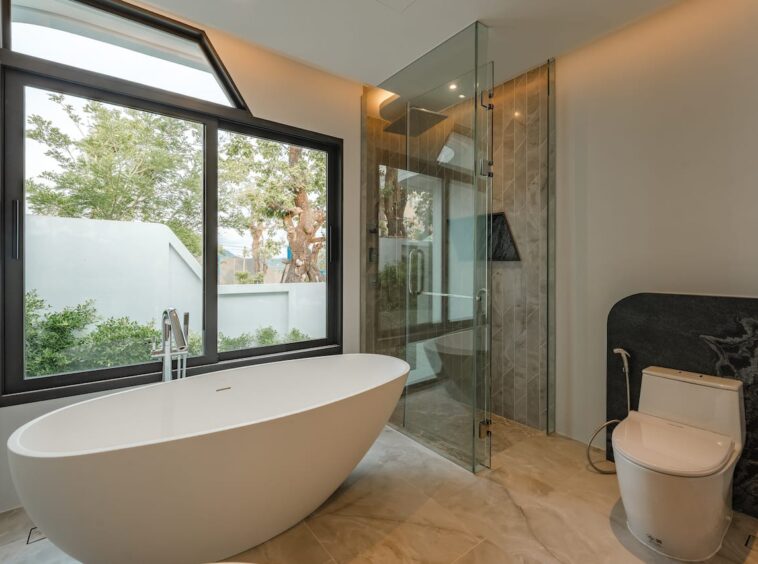 Modern bathroom with a freestanding oval tub by a large window, a glass shower enclosure, and a toilet in a bright, contemporary space.