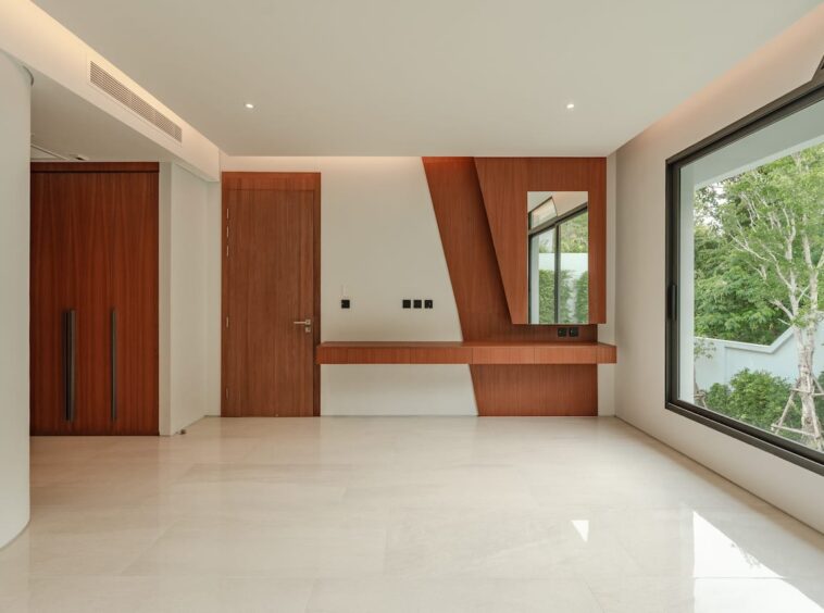 Modern living space with a wooden built-in desk, mirror and large window overlooking greenery outside.
