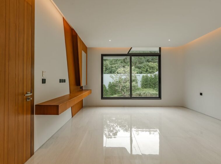 Modern empty room with white glossy floor, wood-paneled wall and floating desk, large window overlooking greenery.